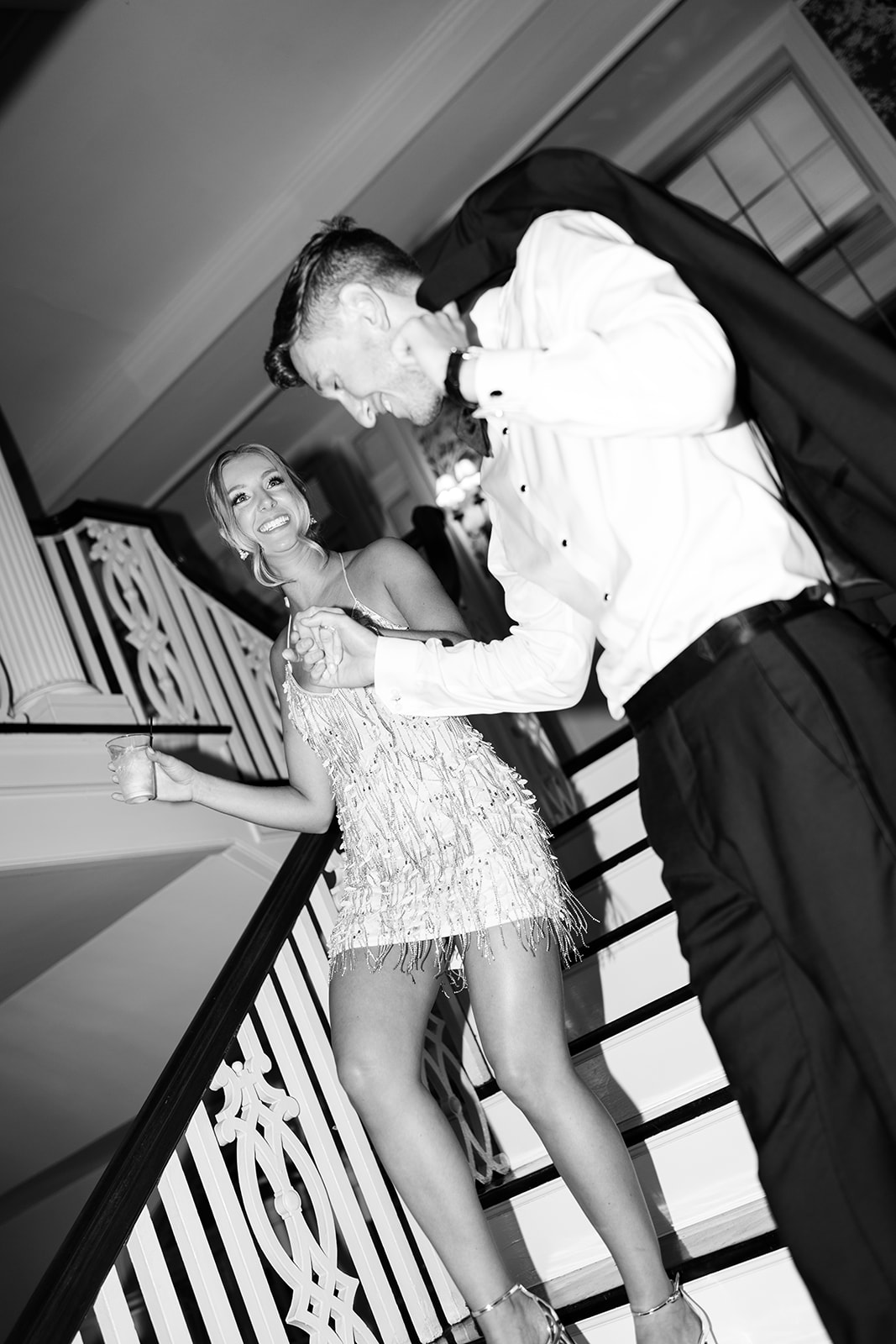 Black and white photo of the bride in a short fringe dress and groom walking down stairs, laughing.